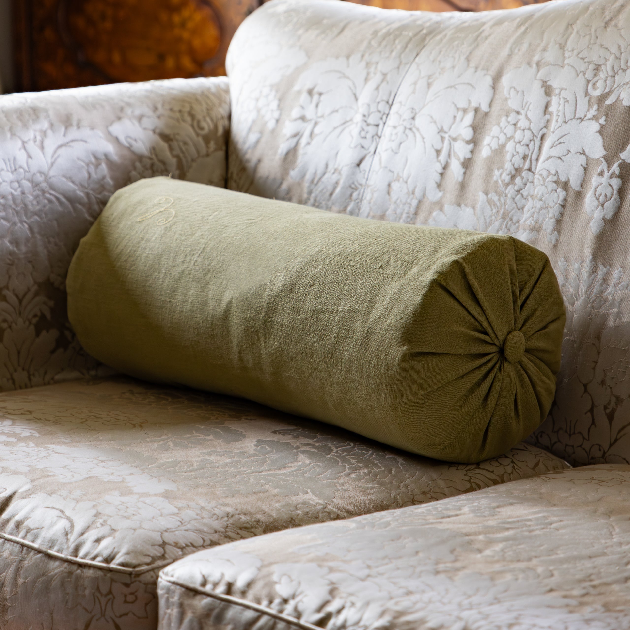 Green cylindrical pillow on a patterned sofa with a wooden cabinet and framed photos in the background.