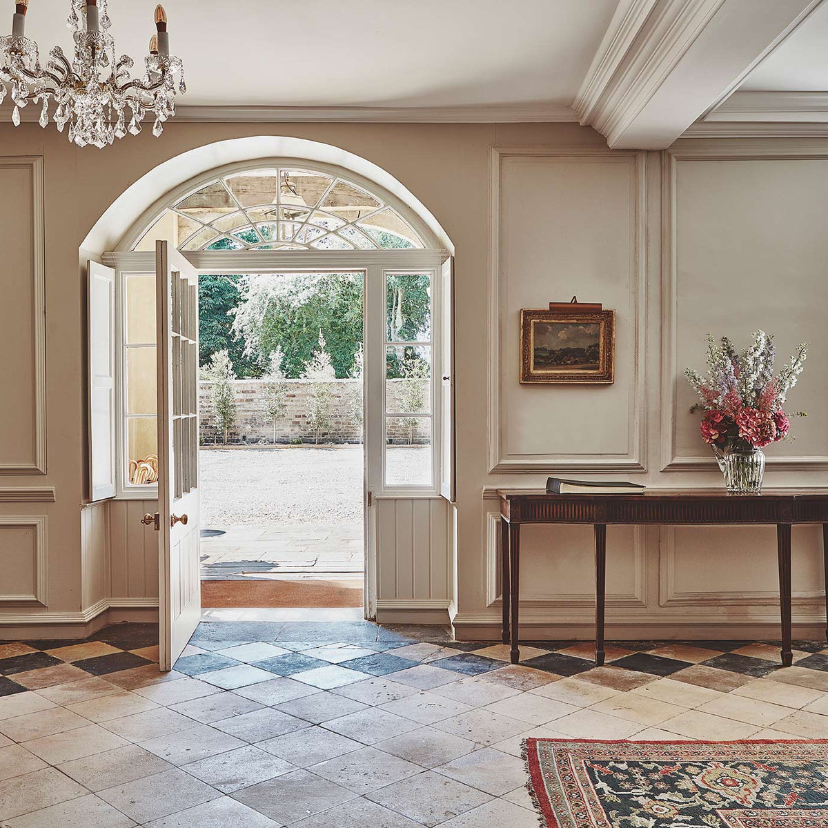 A grand interior room with beige paneled walls, a checkered tile floor, an open arched doorway, a crystal chandelier, and a console table holding a floral arrangement.