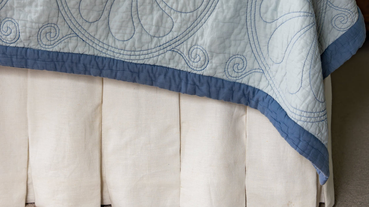 A light blue and white quilted bedspread with floral embroidery and a blue border, styled on a bed with a white skirt and blue pillows in a bedroom setting.