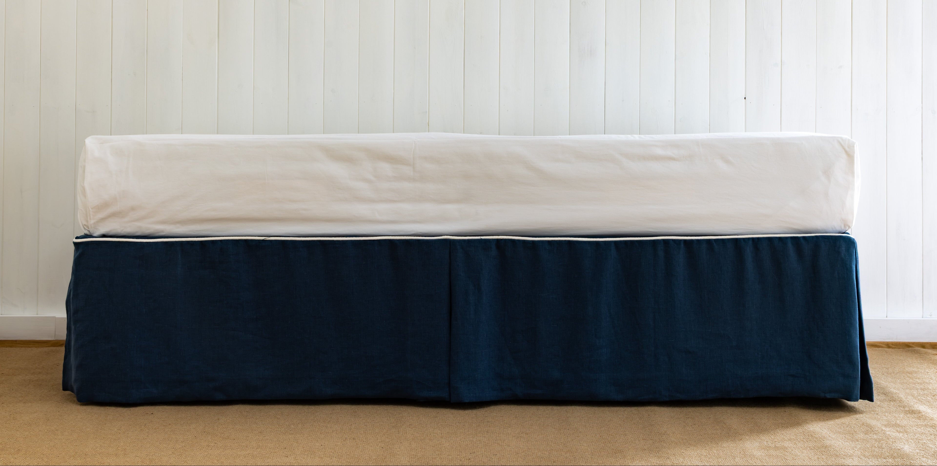Bed with a navy blue bed skirt or bed valance on a wooden floor against a white paneled wall.