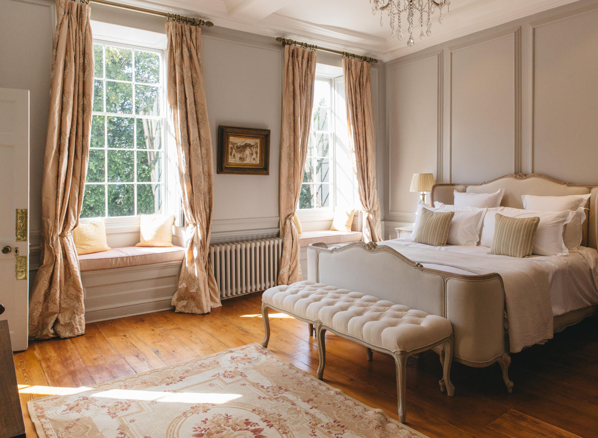 A classic bedroom interior featuring a large bed dressed in white linens, a matching tufted bench at the foot, beige curtains, wood floors, and a patterned rug.